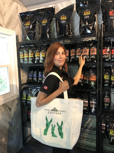 A person holding a white tote bag with the Stone Temple Coffees logo in front of a shelf filled with coffee bags.