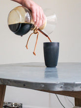 Person pouring coffee from a gold and glass carafe into a black cup on a white countertop.