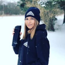 A woman wearing a black beanie with a white coffee logo and holding a black coffee cup, standing outdoors in a snowy environment.